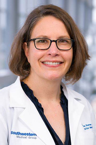 Smiling woman with brown hair wearing a white UT Southwestern Medical Center lab coat over a dark blouse and dark-framed glasses.