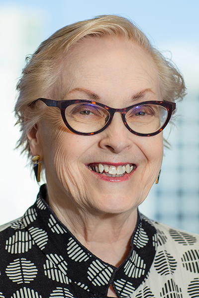 Smiling woman with fair hair wearing a black and gray print blouse and dark-framed glasses.