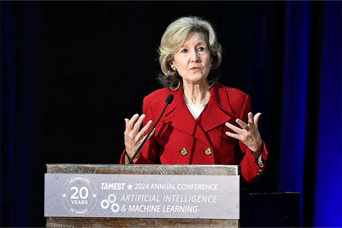 Blond woman wearing a red suit jacket, speaking earnestly to a crowd from a podium. Sign reads - Celebrating 20 years of TAMEST, 2024 Annual Conference, Artificial Intelligence & Machine Learning 