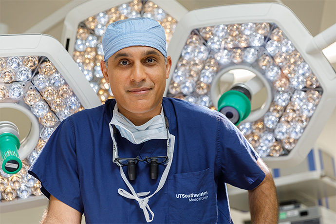 Smiling man wearing blue surgical scrubs with operating room lights and tools behind him.
