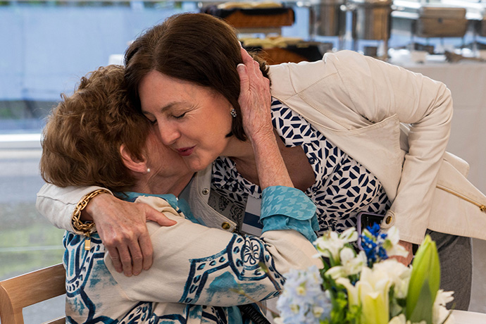 Woman with dark hair wearing a white jacket over a patterned blouse, bends over to hug a woman with short brown hair, seated at a table on which is an arrangement of white and blue flowers.