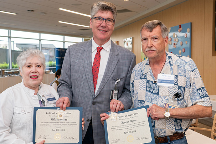 Tall man with graying hair wearing a gray suit with a red tie and dark rimmed glasses, presents Certificates of Appreciation (illegible) to an older couple, Joseph and Hidalia Myers.