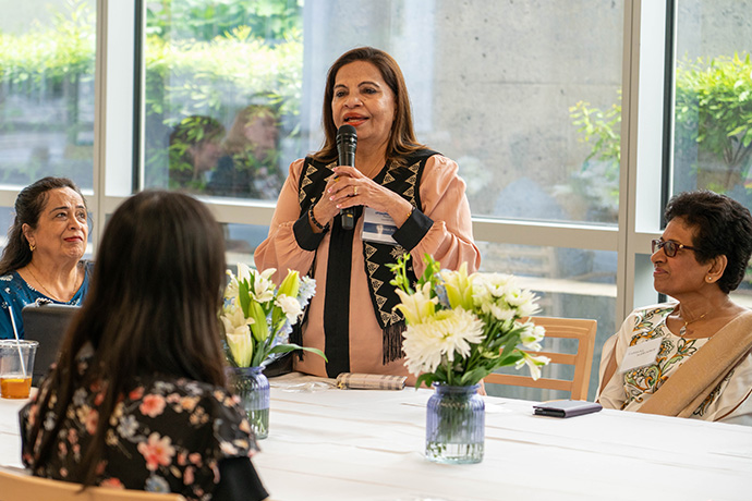 Woman with long dark hair, wearing a beige blouse and black patterned vest, holds a microphone and speaks to a group of women seated at a table on which are two jars of white flowers.