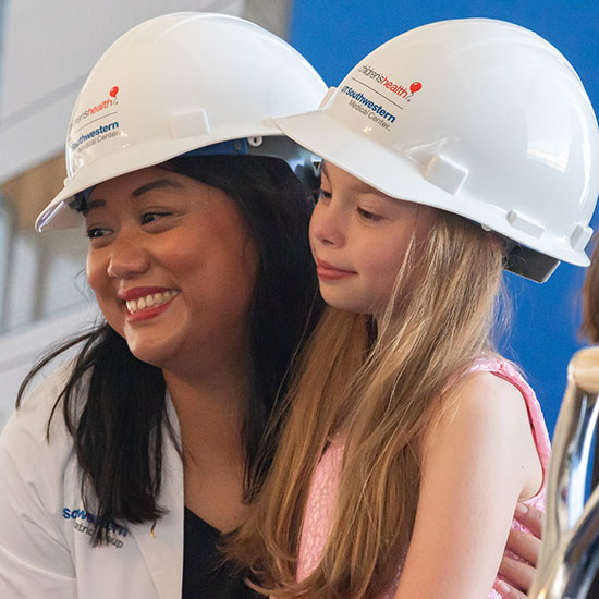 smiling Asian woman in white coat and hard hat puts arm around little blonde girl in pink top and white hard hat