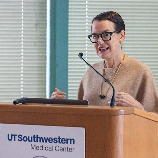 Woman with dark hair and dark rimmed glasses, wearing a beige sweater speaks from a podium bearing the UT Southwestern Medical Center logo.