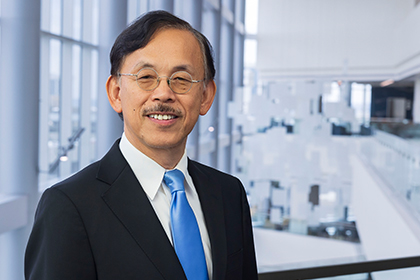 Smiling man dark graying hair and mustache, wearing a dark suit, white shirt, and blue tie.