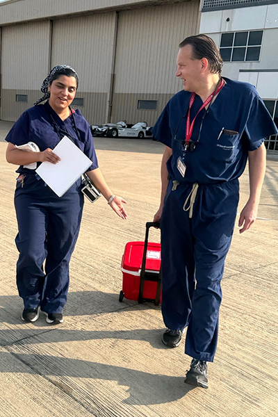 A man and woman in blue scrubs, pulling a red cooler, walk across the airport tarmack.