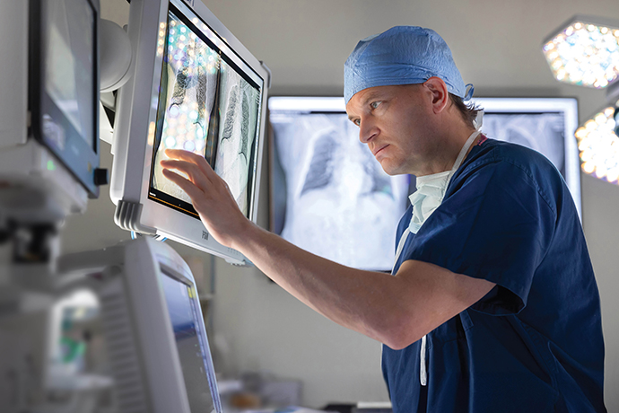 A doctor in blue surgical scrubs examines an X-ray on a wall mounted screen.