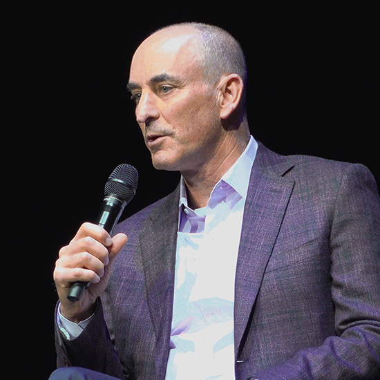 bald white man in grey suit holds microphone as he speaks