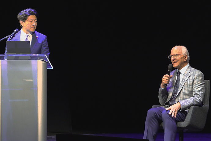 at left stands Asian man in blue suit behind podium looking at balding white man seated to right holding microphone