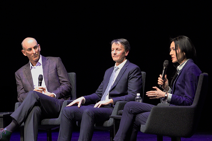 3 people sit in chairs during panel discussion; from left: bald man with microphone, middle-aged man in blue suit and tie, woman with dark hair speaking into microphone