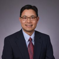 John Zhong, M.D., Asian man, wearing suit, smiling, posing in front of black background