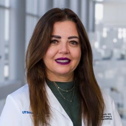 woman with long brown hair, wearing green blouse and white medical coat posed at hospital