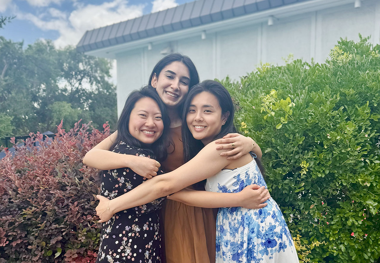 Three women hugging in a green area of campus on a day with blue skies and fluffy clouds