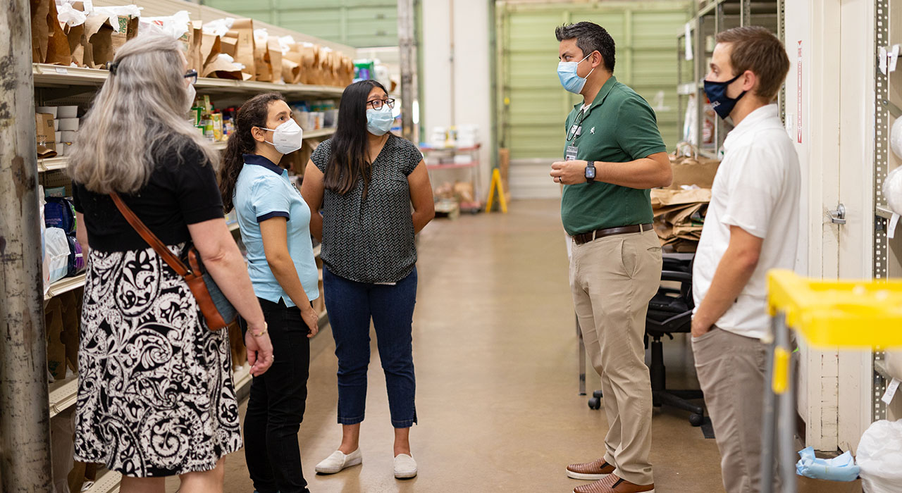 A group of men and women in masks standing in a warehouse that is stocked with food items