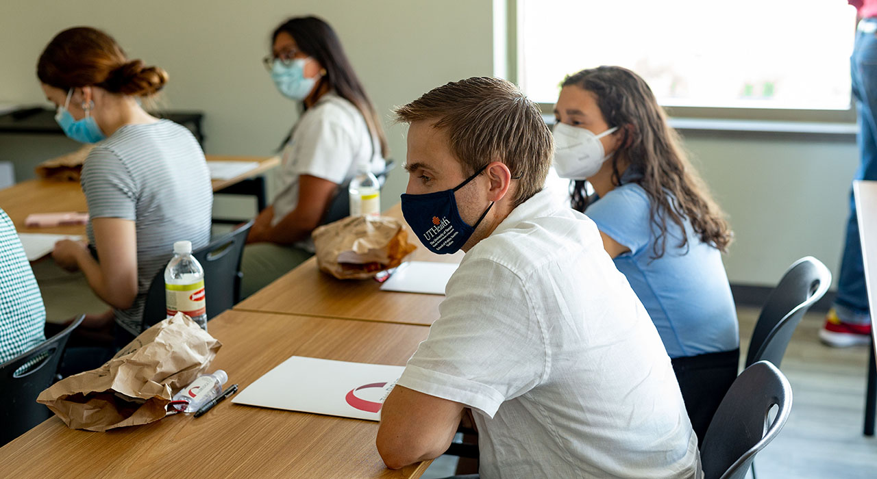 A group of students in masks sitting at desks while watching a presentation