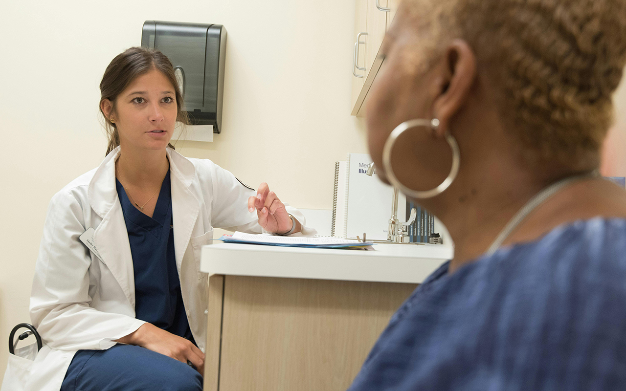 A woman in blue scrubs and white coat speaks with a patient