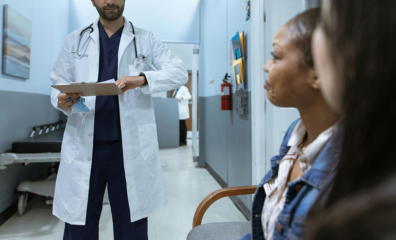 A doctor in blue scrubs and white coat speaks with patient in a clinic
