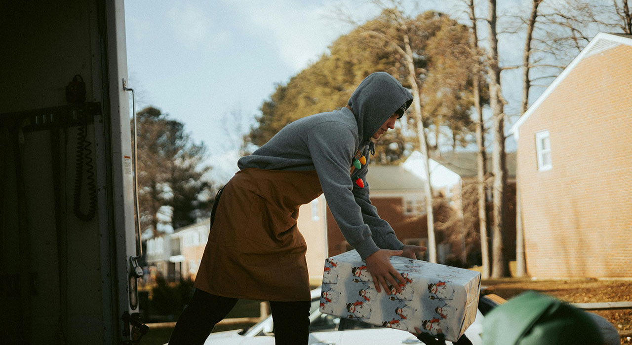 A photo of a person in a grey hoodie and a red apron delivering wrapped presents