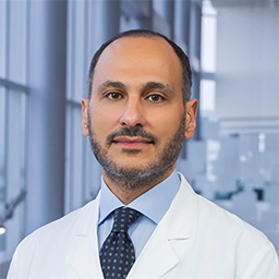 Dr. George Rateb wearing a light blue shirt with a dark patterend tie and a white lab coat standing in the Clements University Hospital lobby