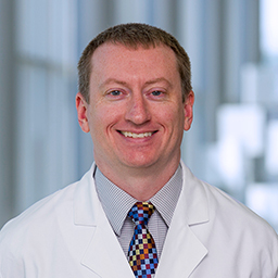 Dr. Markey McNutt wearing  a light blue shirt with a colorful checkered tie and a white lab coat standing in the Clements University Hospital lobby