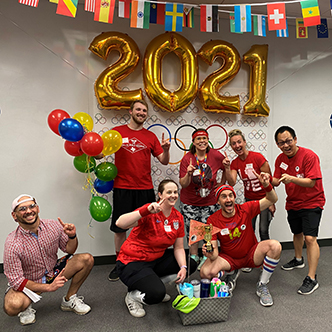 Team of approximately six people pose in front of olympic banner and balloons that spell out 2021