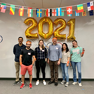 Team of approximately six people pose in front of olympic banner and balloons that spell out 2021
