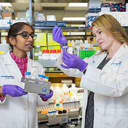 Two women in white lab coats and purple gloves standing in a lab holding vials. One woman is using a dropper to place liquid in a vial as the other woman looks on.