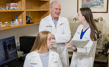 Laughing man in lab looking at woman with long brown hair, with seated woman in the forground who has long blond hair. All are wearing white lab coats.