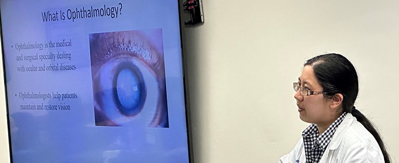 Woman with long dark pony tail, wearing a white lab coat over a black-checked shirt and brown glasses, next to a large screen with copy - What is Ophthalmology? Ophthalmology is the medical and surgical speacialty dealing with ocular and orbital diseases. Ohpthalmologists help patients maintain and restore vision.