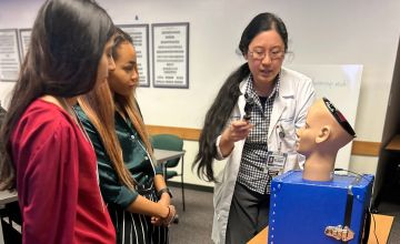 Woman with long dark hair, wearing a white lab coat, holding a tool for eye examination looks at a model of a human head, while two women look on.