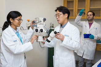 Man and woman, both with black hair and wearing glasses and white lab coats, stand examining a piece of ophthalmic equipment, as a man with black hair wearing glasses and a white lab coat stands in the background holding up a vial of liquid he is examining.. All are wearing white UT Southwestern Medical Center lab coats.