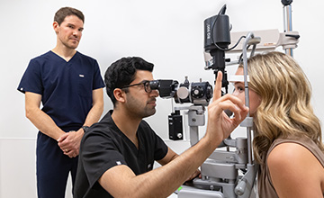 Man with dark hair wearing black scrubs, seated in front of a woman with long blond hair, examining her eyes with ophthalmology machine, while another doctor with dark hair and navy scrubs looks on.