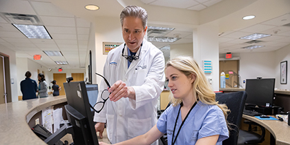 Standing man with gray hair, wearing a white lab coat, using his glasses to point out something on a computer screen to a seated woman with long blond hair, wearing blue scrubs.