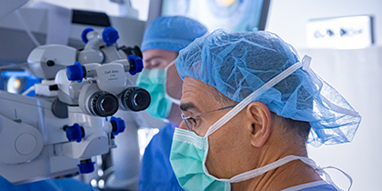 Two men wearing surgical scrubs, caps, and masks, standing with a surgical scope, presumed to be looking at a patient.