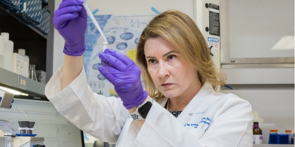 Woman with shoulder length blond hair, wearing a white UT Southwestern lab coat and purple rubber gloves, uses a dropper to place a sample in a tube.