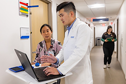 Man with black hair in white lab coat standing in a hallway looking at a laptop while talking to a woman with black hair wearing a black and pink patterned blouse.