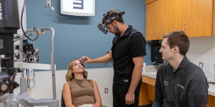 Standing man with dark hair and facial hair, wearing a dark shirt and a testing tool on his head, looks through a lens, down into the eye of a blond, female patient, wearing a brown shirt.