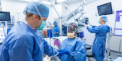 Three individuals in an operating theater, wearing blue scrubs, caps and masks. Individual helping a surgeon on with his gloves is wearing blue scrubs, with a multicolor cap and yellow mask.