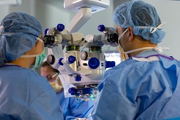 Two individuals dressed in blue scrubs, bonnets, and masks peering into the eye pieces of a piece of large ophthalmic equipment. Their backs are to the camera.