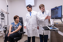 Woman with short brown hair seated in an exam chair looking to her left toward a computer screen. Explaining to her what is on the screen is a man, standing, with black hair and a white lab coat gesturing at the screen while a man (standing) with light brown hair and a white lab coat looks at the screen, too.