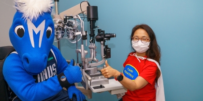 An individual wearing the blue and white Dallas Mavs mascot costume sits oposite a woman with long dark hair, wearing a white mask, red shirt with a blue logo, and white cape. Woman is giving a thumbs-up sign.