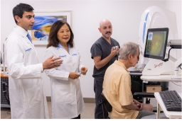 Man, standing, with black hair wearing a white lab coat speaks with a woman with black hair, also wearing a white lab coat. Standing next to them is a man with a bald head wearing grey surgical scrubs operating a piece of ophthalmic imaging equipment on an elderly male patient in a yellow shirt seated at the equipment as his eyes are examined.