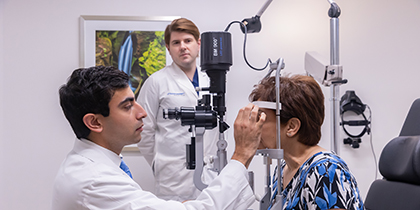 Man with brown hair wearing a white lab coat watches as a seated man with dark hair uses ophthalmology equipment examines the eyes of a woman with short auburn hair, wearing a blue, black, and white print blouse.