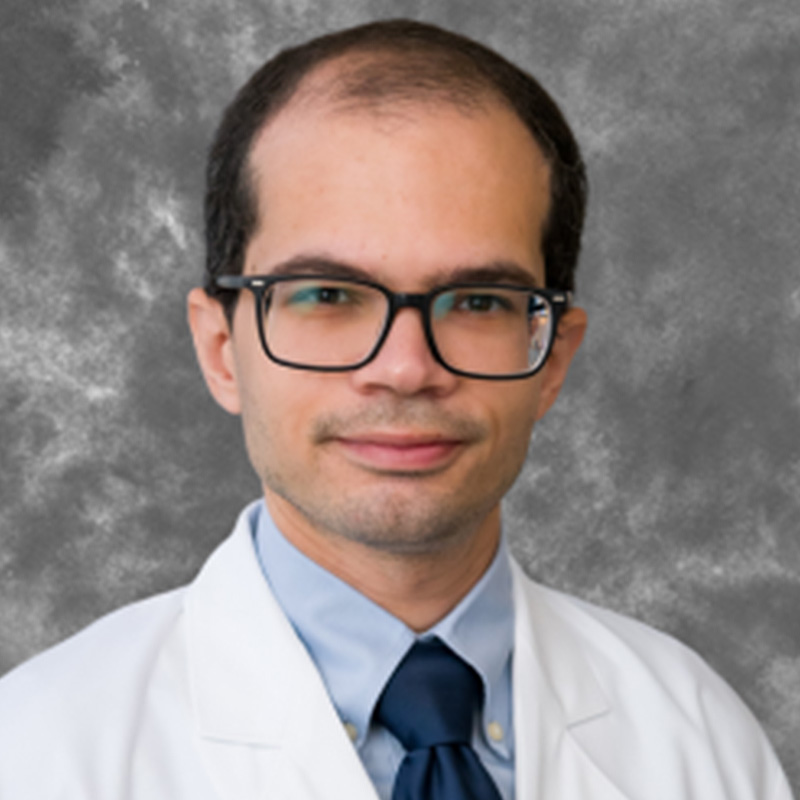 Slightly smiling man with receding dark hair, wearing a white lab coat, blue shirt, navy tie, and dark-rimmed glasses.