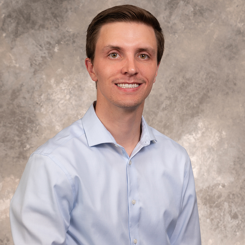 Smiling young man with short brown hair, wearing a dark suit and white dress shirt.