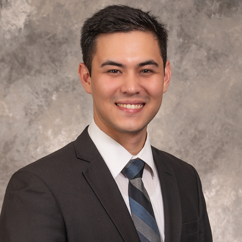 Smiling young man with short dark hair and the start of a dark mustache, wearing a dark suit with white shirt.