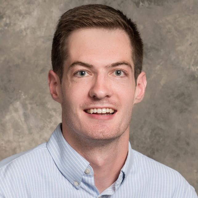 Smiling young man with short brown hair, wearing a blue and white striped dress shirt.