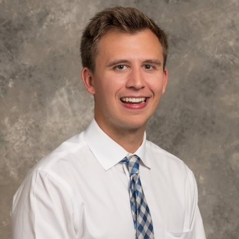 Smiling young man with brown hair, wearing a white dress shirt and blue plaid tie.