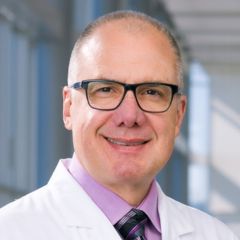 Smiling man with receding gray hair, wearing a purple dress shirt, dark tie, and dark-rimmed glasses.